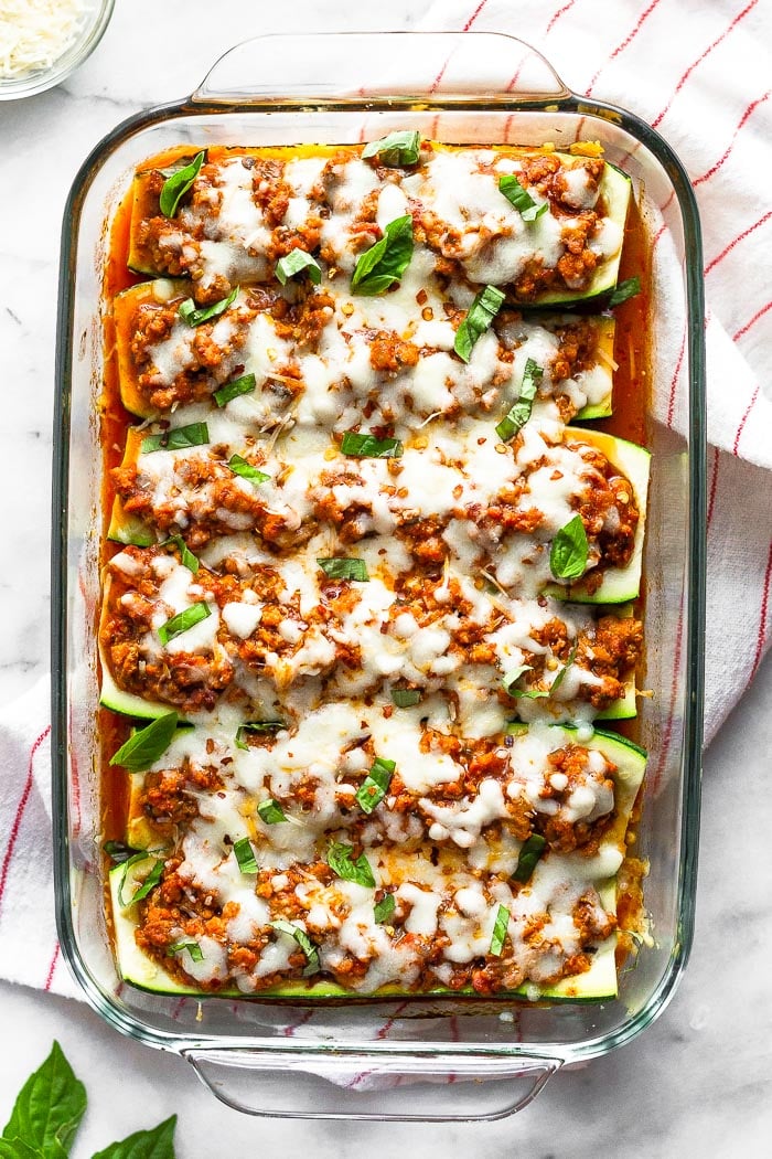 Large glass baking dish filled with healthy zucchini pizza boats with sausage. The dish is sitting on towel and around it is fresh basil and small bowl of parmesan cheese.