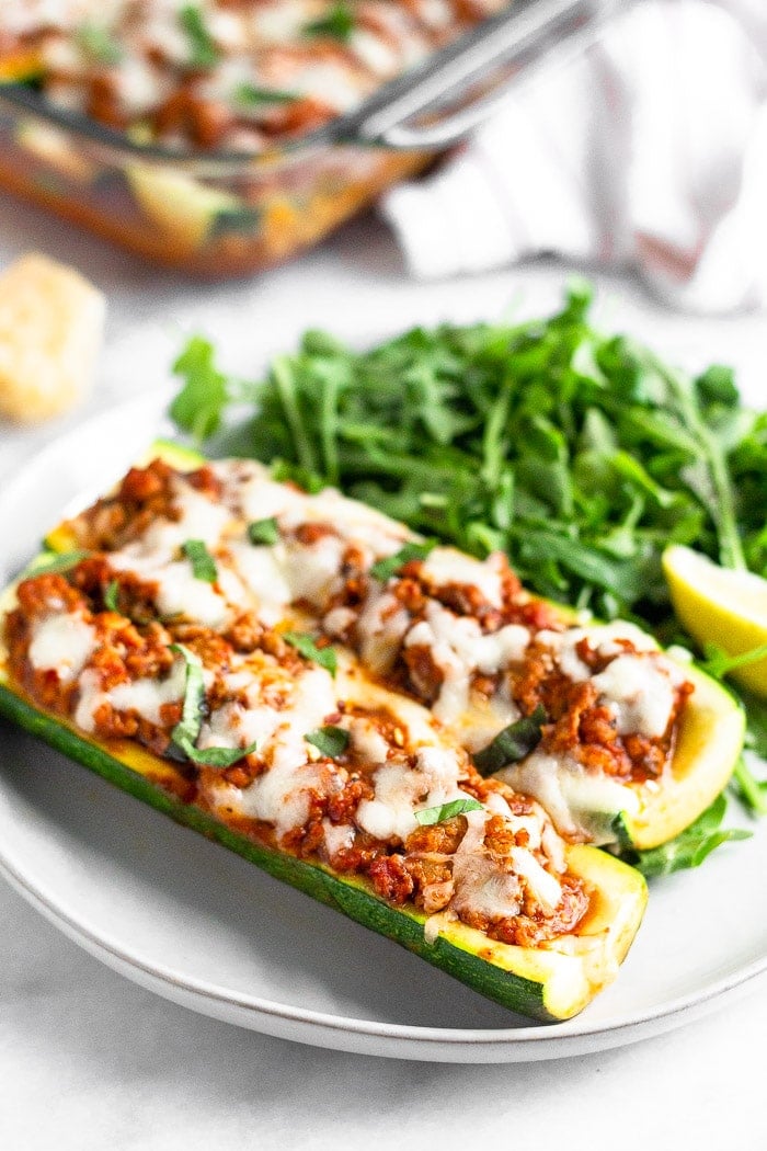 Two keto zucchini pizza boats with sausage on a plate with greens and a lemon wedge. Behind it is block or parmesan cheese, a pan of more zucchini pizza boats, and a kitchen towel.