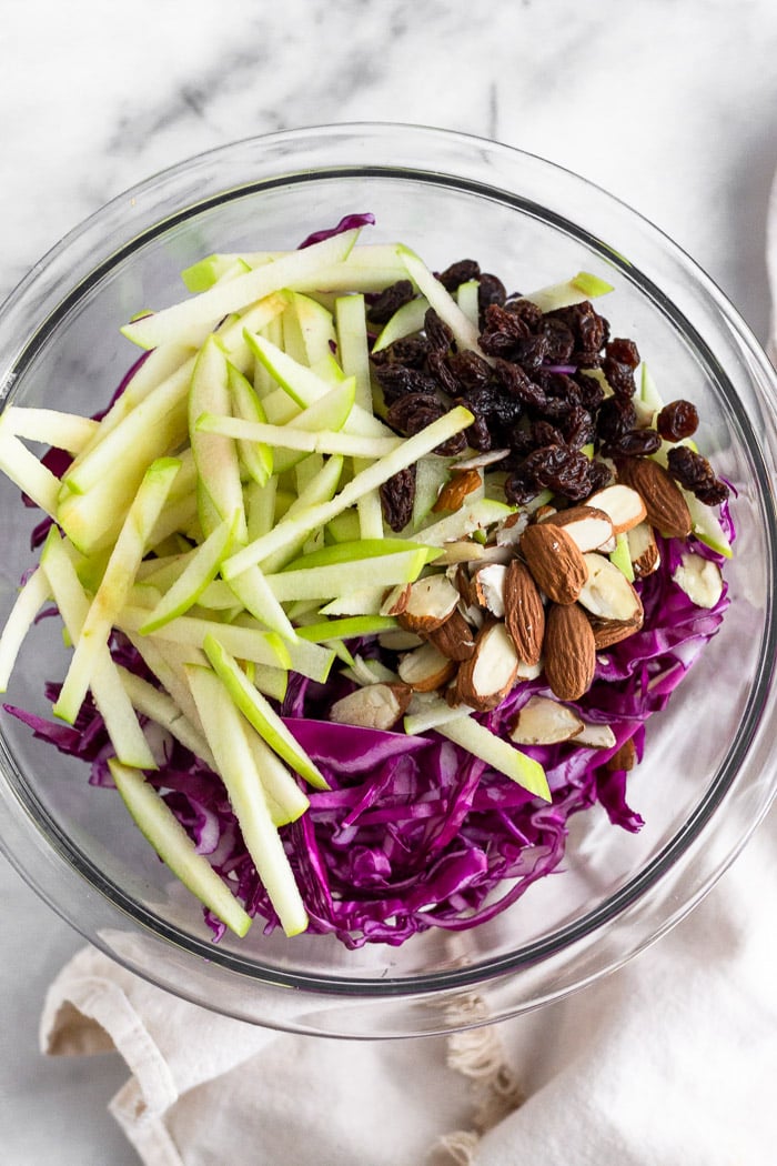 Bowl of shredded cabbage, apples cut into matchsticks, raisins, and chopped almonds.