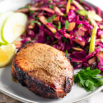 Plate with air fryer pork chops, cabbage apple salad, sliced apples, a lemon wedge, and fresh parley. It is on a towel and behind it is another plate.