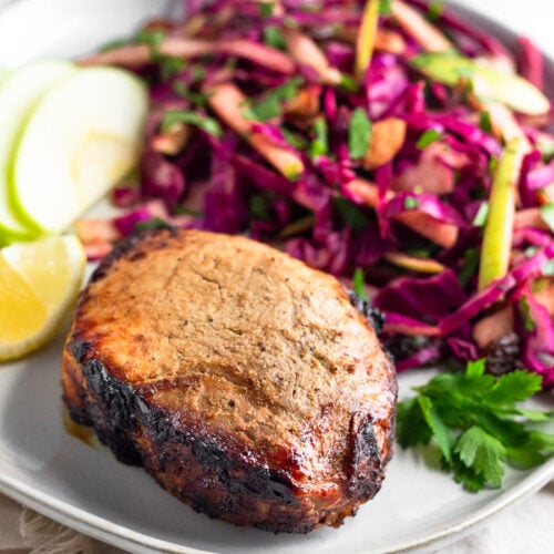 Plate with air fryer pork chops, cabbage apple salad, sliced apples, a lemon wedge, and fresh parley. It is on a towel and behind it is another plate.