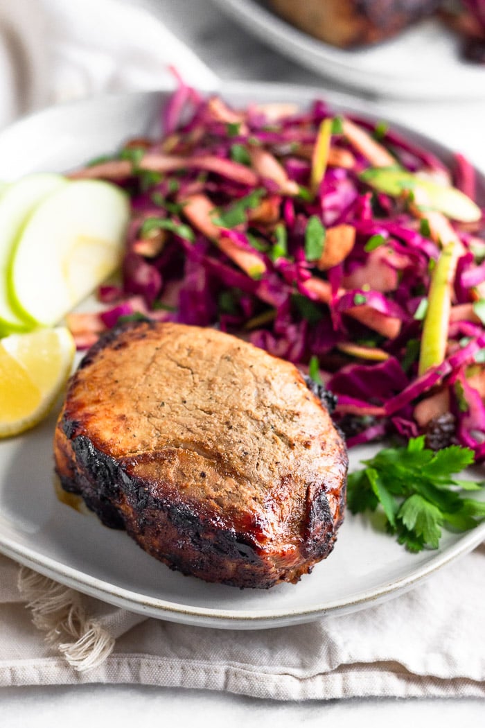 Plate with air fryer pork chops, cabbage apple salad, sliced apples, a lemon wedge, and fresh parley. It is on a towel and behind it is another plate.