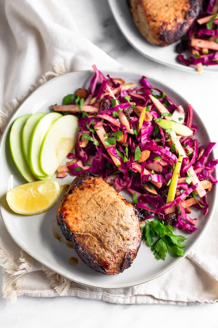 Overhead of a plate with air fryer pork chops, cabbage apple salad, apple slices, and a lemon wedge. It is on a kitchen towel with another plate next with another pork chop and more salad on it.