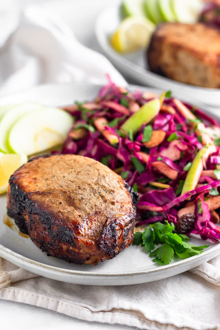 White plate sitting on a white towel with air fryer pork chops, cabbage apple salad, sliced apples, a lemon wedge, and fresh parley. Behind it is another plate with the same things on it.