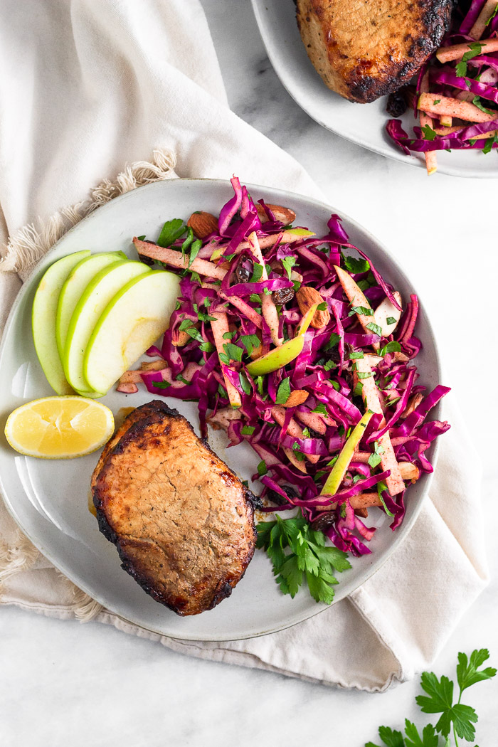Overhead of a white plate with air fryer pork chops, cabbage salad, apple slices, and a lemon wedge. It is on a kitchen towel with another plate next to it and some fresh parsley.