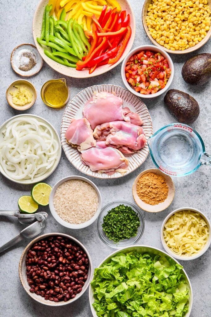 Grey concrete counter with a bowl of corn, a bowl of pico de gallo, two whole avocados, a measuring cup of water, a small bowl of taco seasoning, a small bowl of chopped cilantro, a bowl of shredded cheese, a bowl of chopped lettuce, a bowl of black beans, a bowl of uncooked white rice, a citrus press, a lime that has been cut in half, a bowl of raw chicken thighs, a bowl of sliced white onions, a small cup of olive oil, a small bowl of minced garlic, a small bowl of salt and pepper, and a bowl of sliced red, green, and yellow bell peppers on it.
