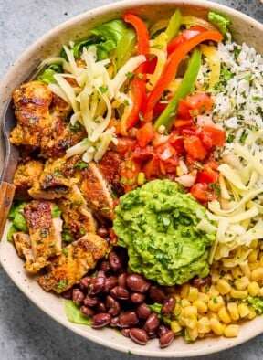 Overhead shot of a chicken burrito bowl with cilantro lime rice, corn, black beans, guacamole, sliced seasoned chicken, lettuce, shredded cheese, fajita veggies, and pico de gallo. A fork is in the bowl as well.