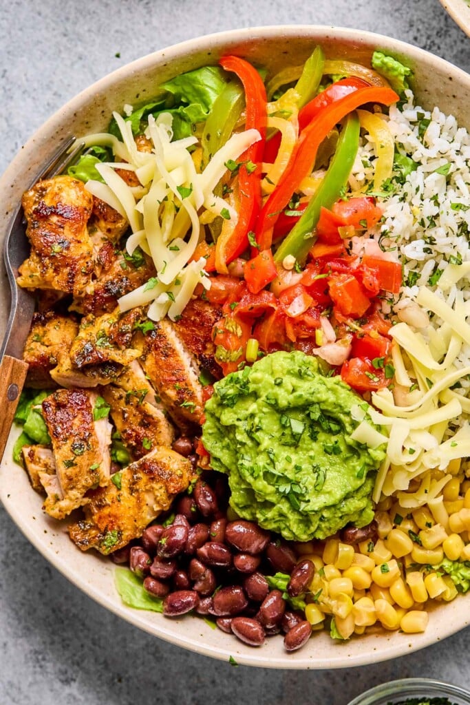 Overhead shot of a chicken burrito bowl with cilantro lime rice, corn, black beans, guacamole, sliced seasoned chicken, lettuce, shredded cheese, fajita veggies, and pico de gallo. A fork is in the bowl as well.