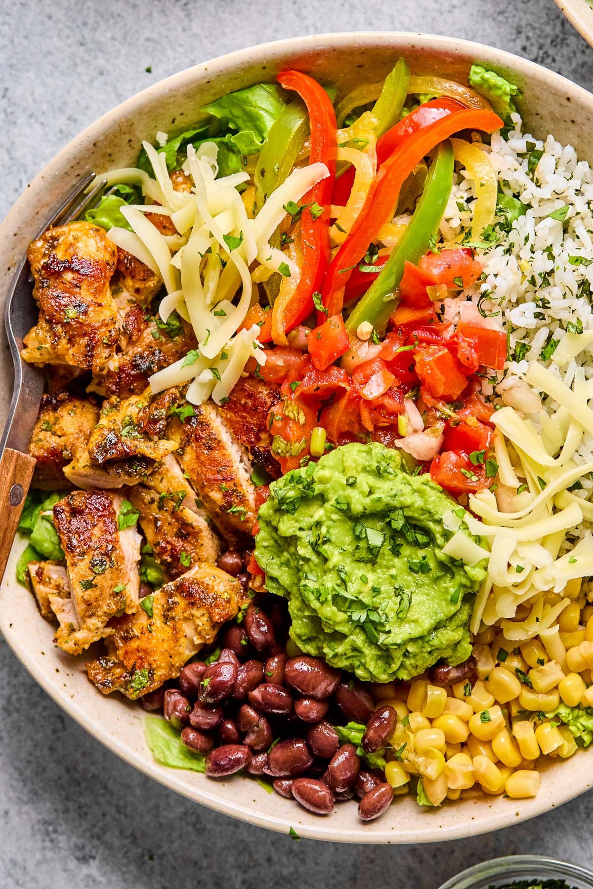 Overhead shot of a chicken burrito bowl with cilantro lime rice, corn, black beans, guacamole, sliced seasoned chicken, lettuce, shredded cheese, fajita veggies, and pico de gallo. A fork is in the bowl as well.