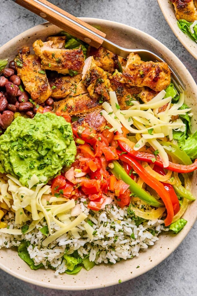 Overhead shot of a white bowl with seasoned sliced chicken, cilantro lime rice, sautéed onions and peppers, black beans, lettuce, shredded cheese, salsa, and guacamole. In the bowl is a fork.