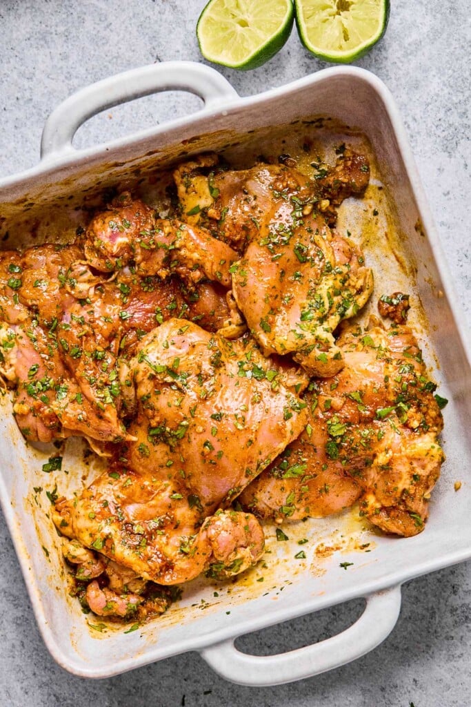 Overhead shot of raw chicken thighs in a baking dish coated with oil, lime juice, cilantro, and spices. Next to the baking dish are two halves of limes that have been juiced.