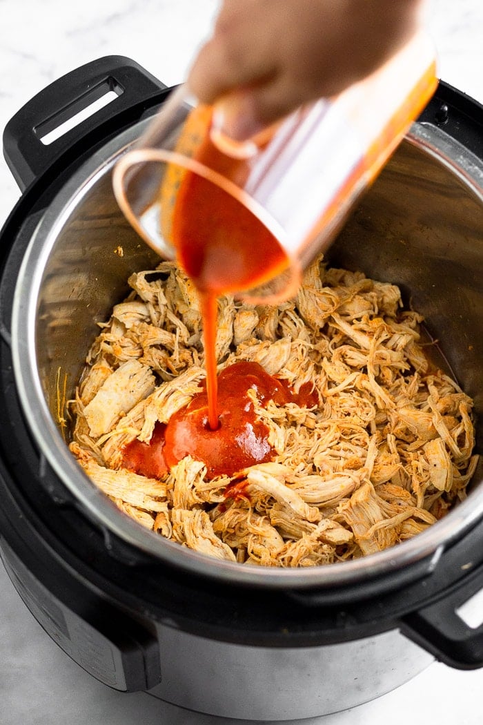 Pressure cooker filled with shredded chicken with a measuring cup pouring buffalo sauce in it.