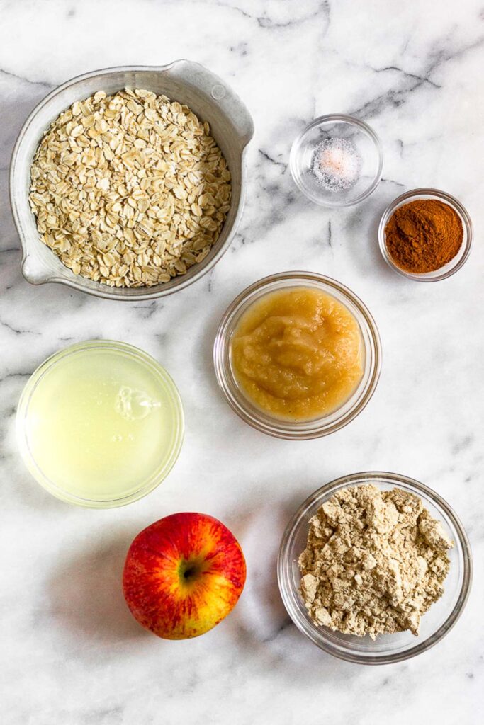 A white marble counter with a small bowl of salt, a small bowl of cinnamon, a bowl of applesauce, a bowl of vanilla protein powder, a red apple, a bowl of egg whites, and a bowl of rolled oats.