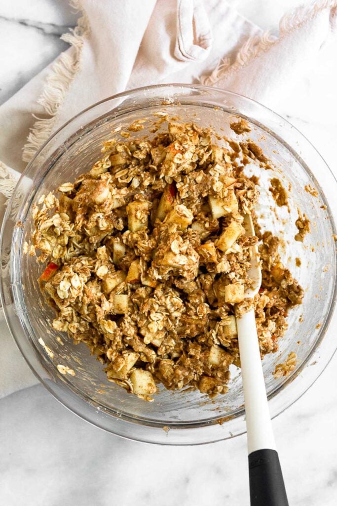 A bowl filled with a wet mixture of oats and diced apple with a spatula in it. The bowl is sitting on a white kitchen towel.