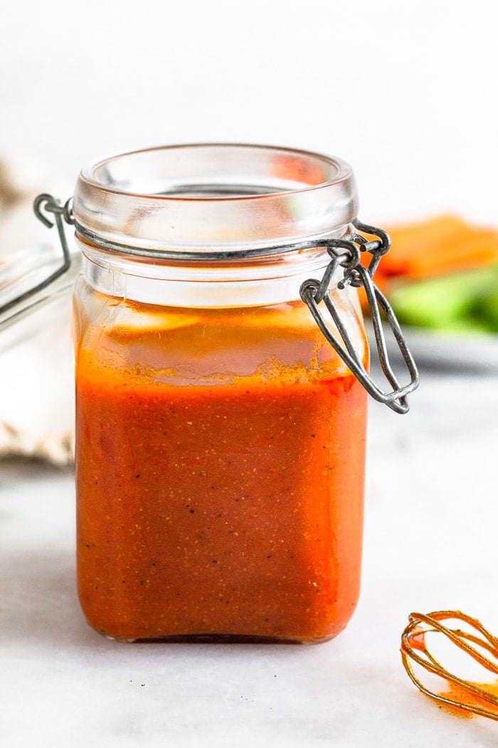 Jar filled with healthy buffalo sauce with a whisk in front of it and plate of veggies behind it.