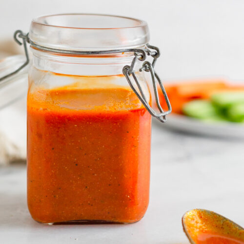 Small jar of homemade Whole30 buffalo sauce recipe. In front of it is a spoon with sauce on it and behind it is a plate of carrots and celery.