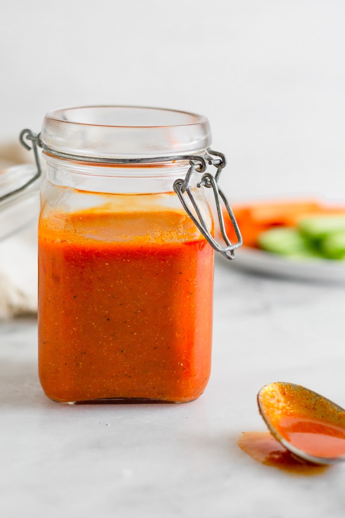 Small jar of homemade Whole30 buffalo sauce recipe. In front of it is a spoon with sauce on it and behind it is a plate of carrots and celery.