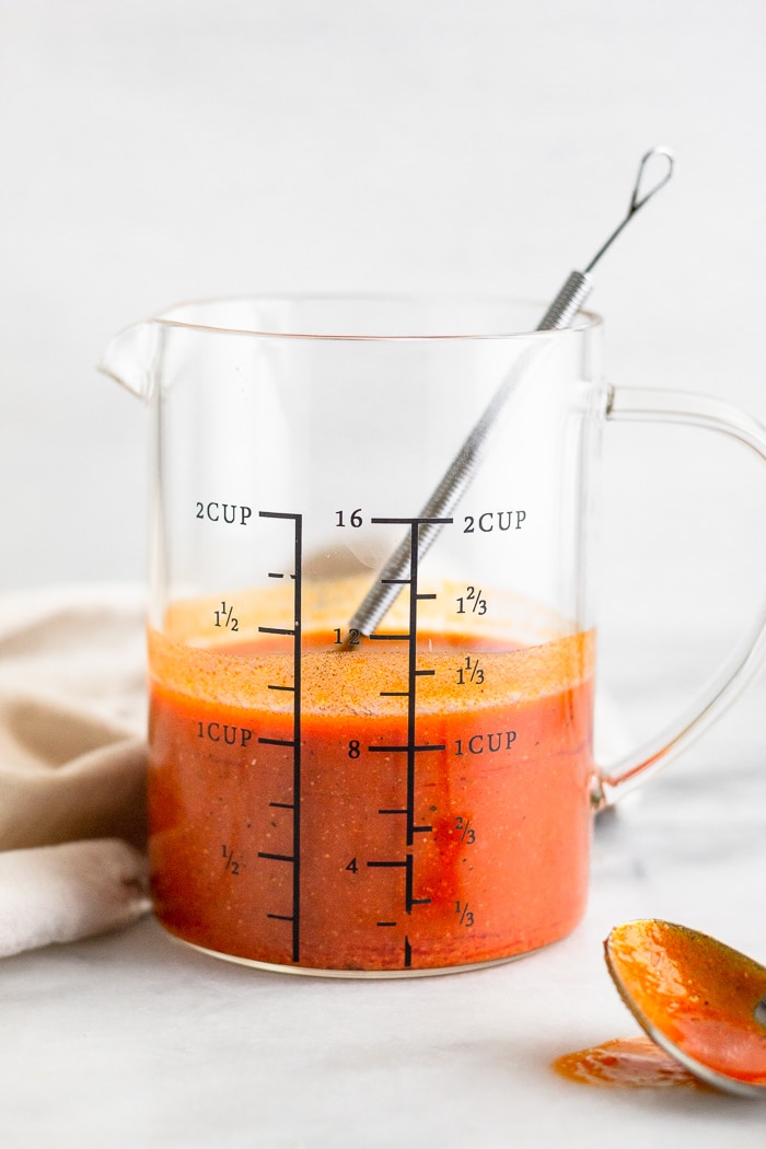 Measuring cup filled with the best buffalo sauce. A small whisk in the cup with a spoon in front of them.