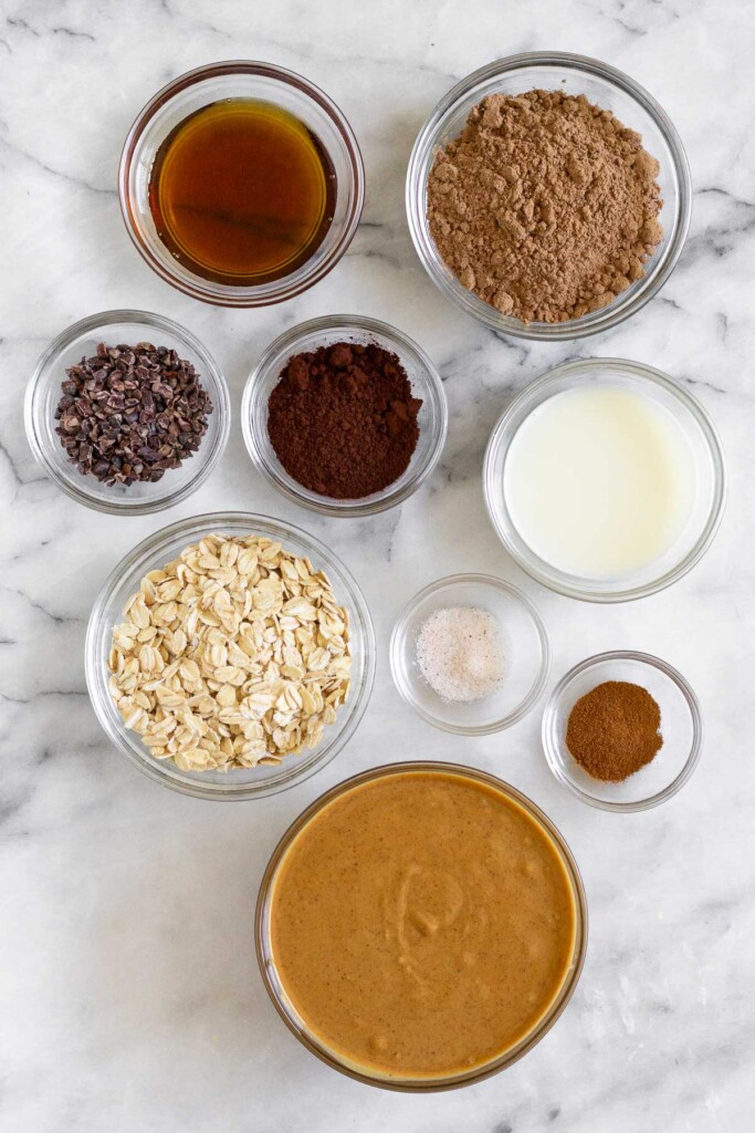 White marble counter with a bowl of chocolate protein powder, a bowl of milk, a small bowl of salt, a small bowl of cinnamon, a bowl of creamy peanut butter, a bowl of rolled oats, a bowl of cocoa powder, a bowl of cacao nibs, and a bowl of maple syrup.