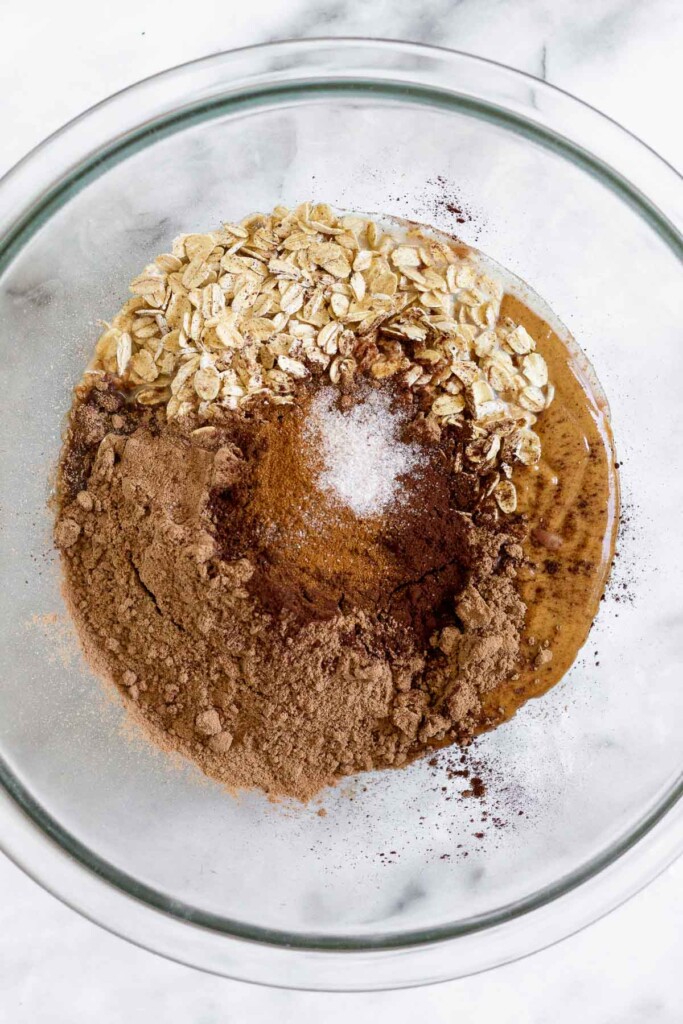 A glass bowl with rolled oats, nut butter, chocolate protein powder, cocoa powder, cinnamon, and salt before it is mixed.