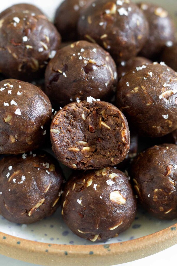 A close up of a pile of chocolate peanut butter energy balls sprinkled with flaky sea salt. The one in the front as a bite taken out of it.