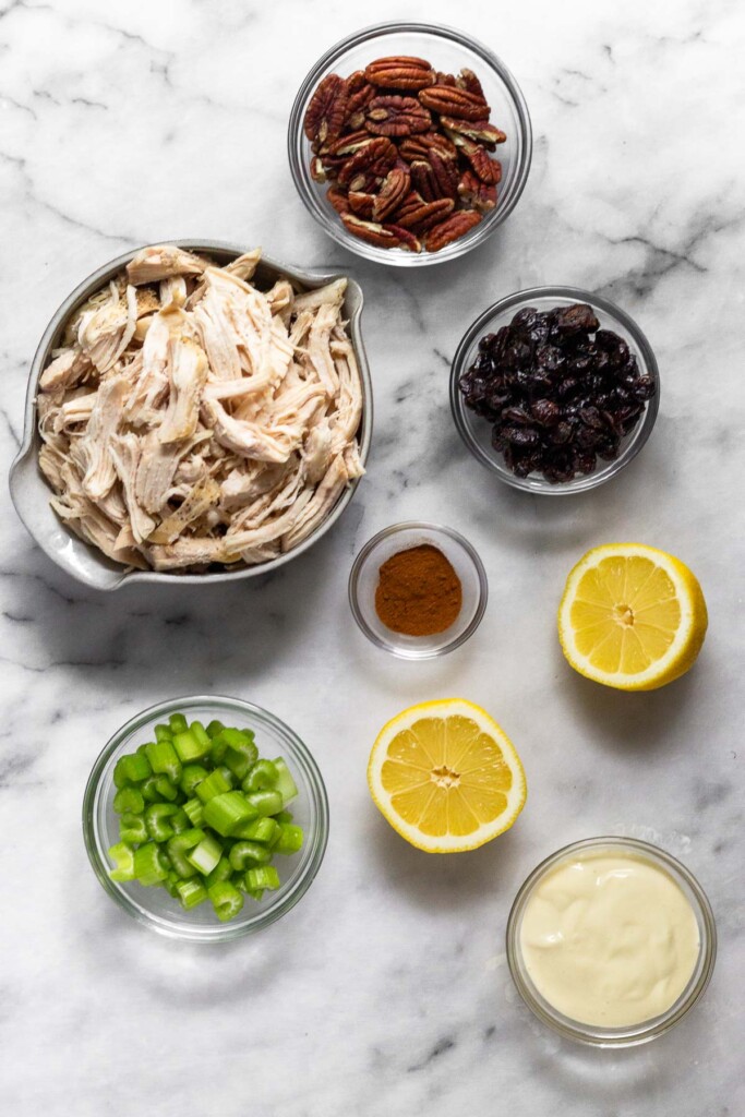 White marble counter with a bowl of pecans, a bowl of dried cranberries, a bowl of cinnamon, a lemon cut in half, a bowl of mayo, a bowl of diced celery, and a bowl of shredded chicken.