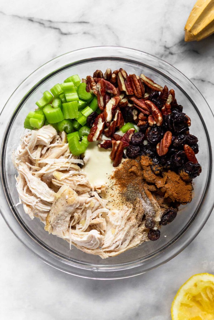 A large glass bowl shredded chicken, diced celery, chopped pecans, dried cranberries, mayo, cinnamon, salt, and pepper.