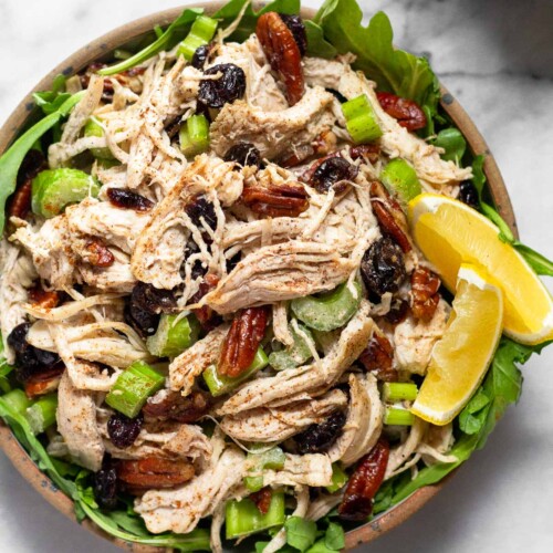 Cranberry pecan chicken salad overtop some greens in a bowl and garnished with 2 lemon wedges. Around the bowl is a plate of celery and carrots and a bowl of pecans.