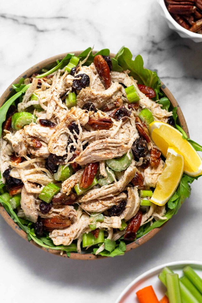 Cranberry pecan chicken salad overtop some greens in a bowl and garnished with 2 lemon wedges. Around the bowl is a plate of celery and carrots and a bowl of pecans.