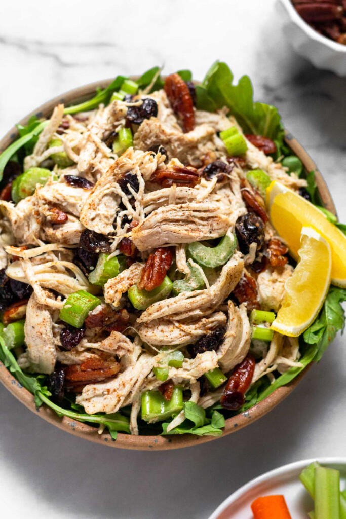 Cranberry chicken salad with pecans and celery in a bowl. It is overtop some mixed greens and garnished with 2 lemon wedges.