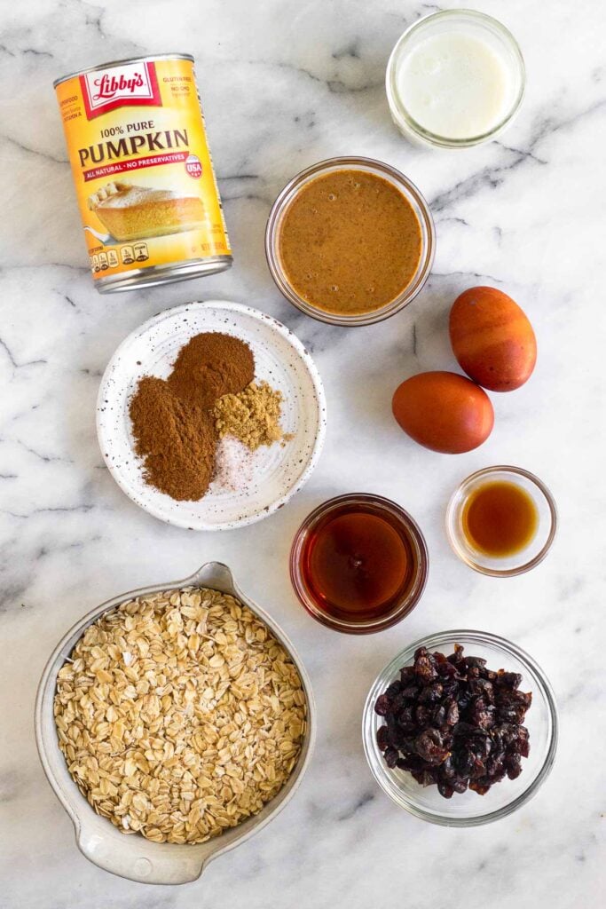 White counter with a can of pumpkin puree, a bowl of almond butter, a jar of milk, 2 eggs, a bowl of vanilla extract, a bowl of maple syrup, a bowl of dried cranberries, a bowl of oats, and a plate of spices.