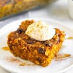 A piece of pumpkin baked oatmeal on a small white plate topped with greek yogurt, cinnamon, and honey. Behind it is a pan of more baked oatmeal.