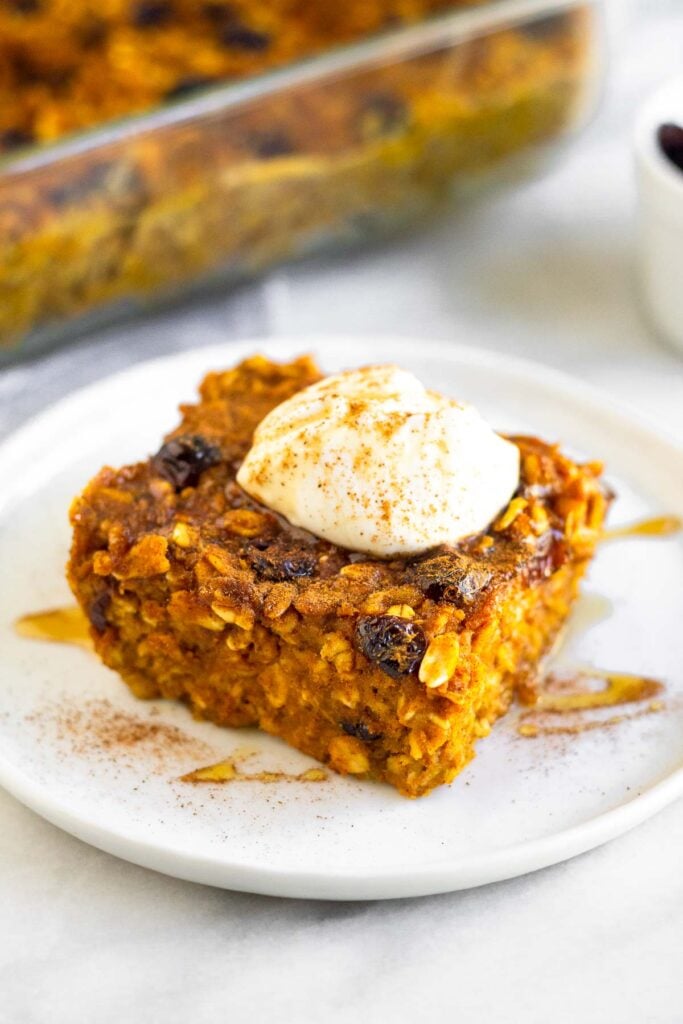 A piece of pumpkin baked oatmeal on a small white plate topped with greek yogurt, cinnamon, and honey. Behind it is a pan of more baked oatmeal.