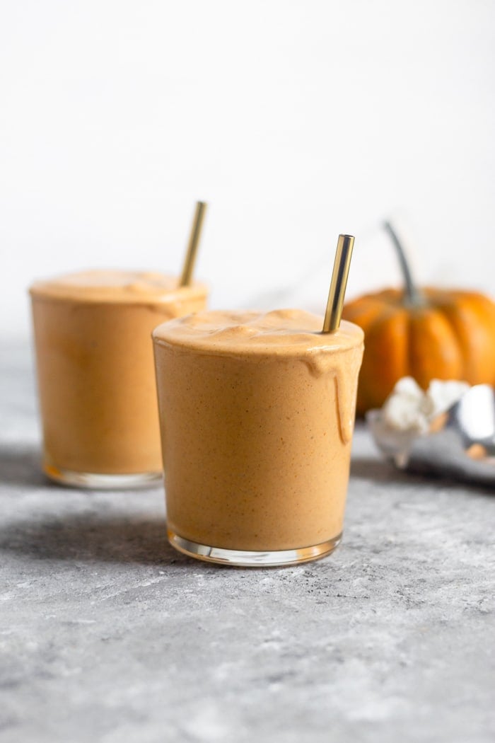 Two pumpkin vegan milkshakes in glass cups with straws in them. Behind them is an ice cream scoop with ice cream in it and a pumpkin.