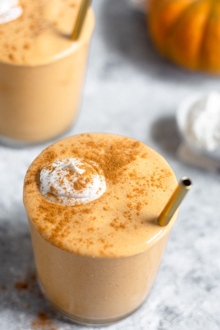 Overhead shot of a pumpkin spice milkshake topped with whipped cream and cinnamon with a straw coming out. Behind it is another glass, a pumpkin, and an ice cream scoop filled with vanilla ice cream.