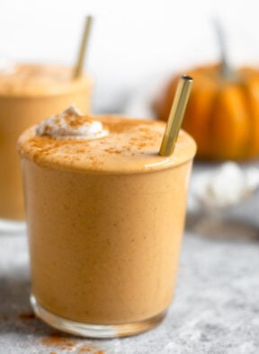 Close up of a pumpkin milkshake in a glass cup. It is topped with whipped cream and cinnamon with a straw sticking out of it. Behind it is another cup and a pumpkin.