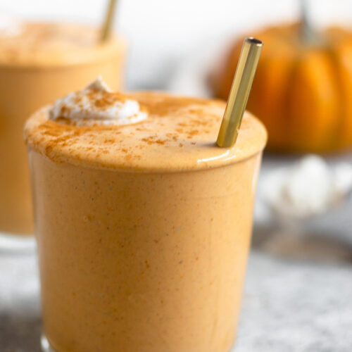 Close up of a pumpkin milkshake in a glass cup. It is topped with whipped cream and cinnamon with a straw sticking out of it. Behind it is another cup and a pumpkin.