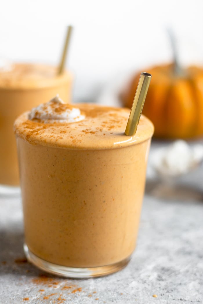 Close up of a pumpkin milkshake in a glass cup. It is topped with whipped cream and cinnamon with a straw sticking out of it. Behind it is another cup and a pumpkin.