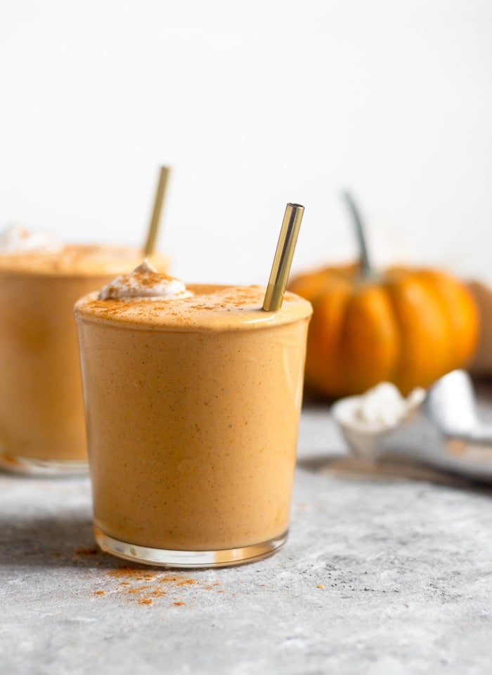 Pumpkin pie milkshake in a glass cup topped with whipped cream and cinnamon with a straw in it. Behind it is another cup along with a pumpkin and an ice cream scoop filled with ice cream.