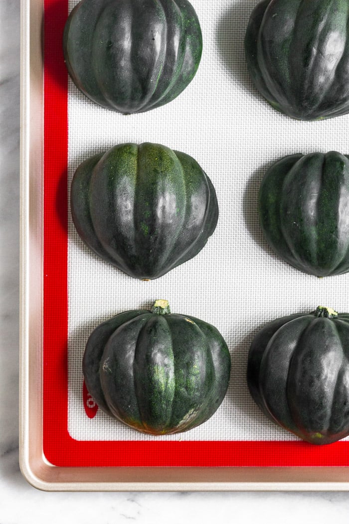 Halved acorn squash sitting on a silicone lined baking sheet that flesh side down.