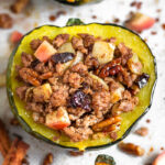 Sausage stuffed acorn squash filled with ground pork, apples, pecans, cranberries, and spices. It is sitting on a baking sheet surrounded by more stuffed squash with the filling scattered across on the silicone mat they are sitting on.