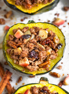 Sausage stuffed acorn squash filled with ground pork, apples, pecans, cranberries, and spices. It is sitting on a baking sheet surrounded by more stuffed squash with the filling scattered across on the silicone mat they are sitting on.