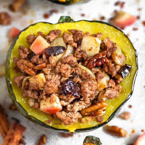 Sausage stuffed acorn squash filled with ground pork, apples, pecans, cranberries, and spices. It is sitting on a baking sheet surrounded by more stuffed squash with the filling scattered across on the silicone mat they are sitting on.