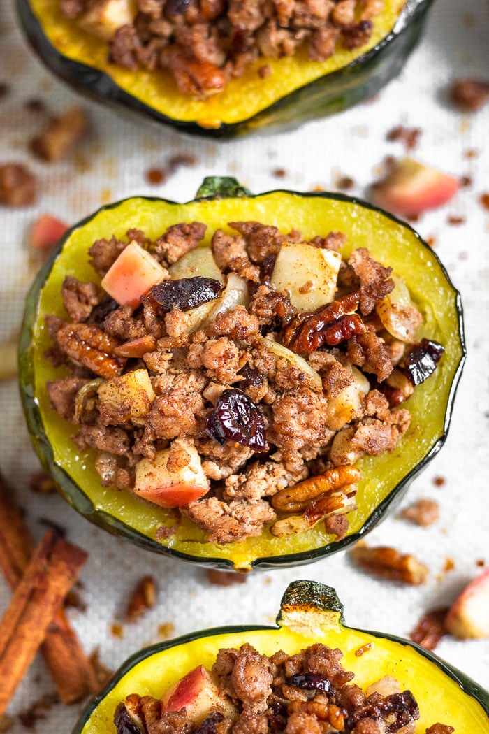 Sausage stuffed acorn squash filled with ground pork, apples, pecans, cranberries, and spices. It is sitting on a baking sheet surrounded by more stuffed squash with the filling scattered across on the silicone mat they are sitting on.