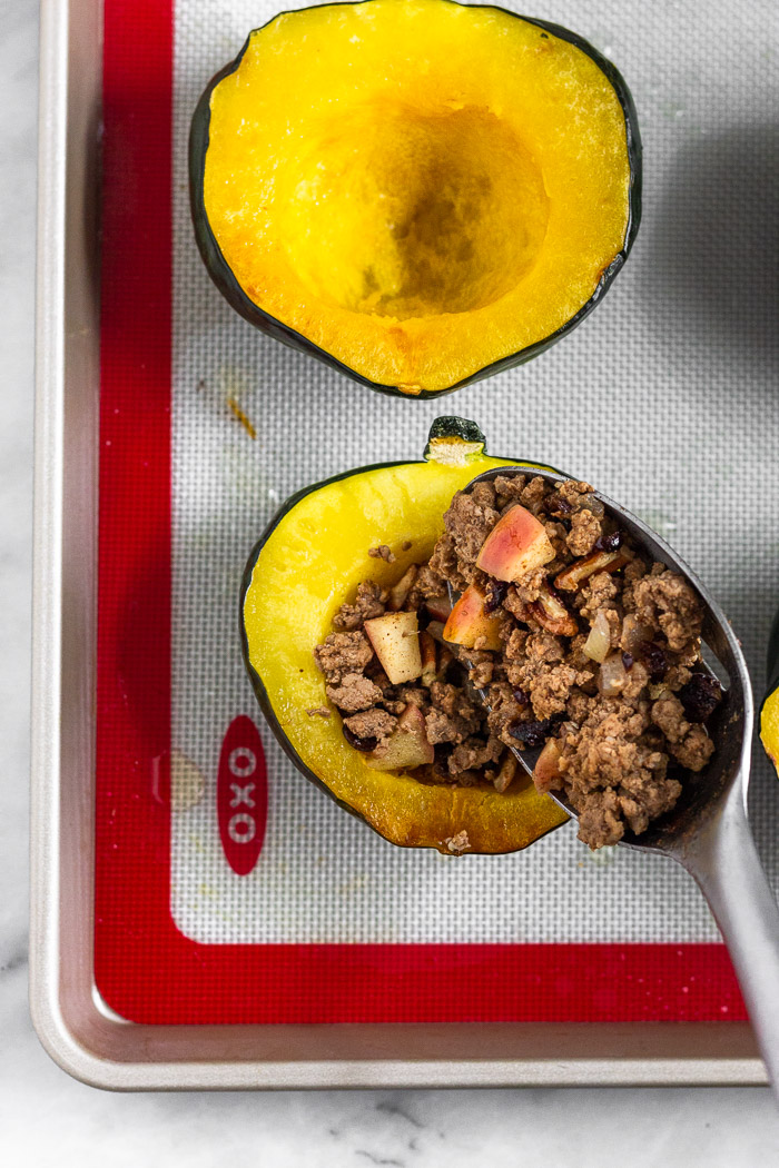 Roasted acorn squash on baking sheet that is being filled with a cooked sausage and apple mixture.