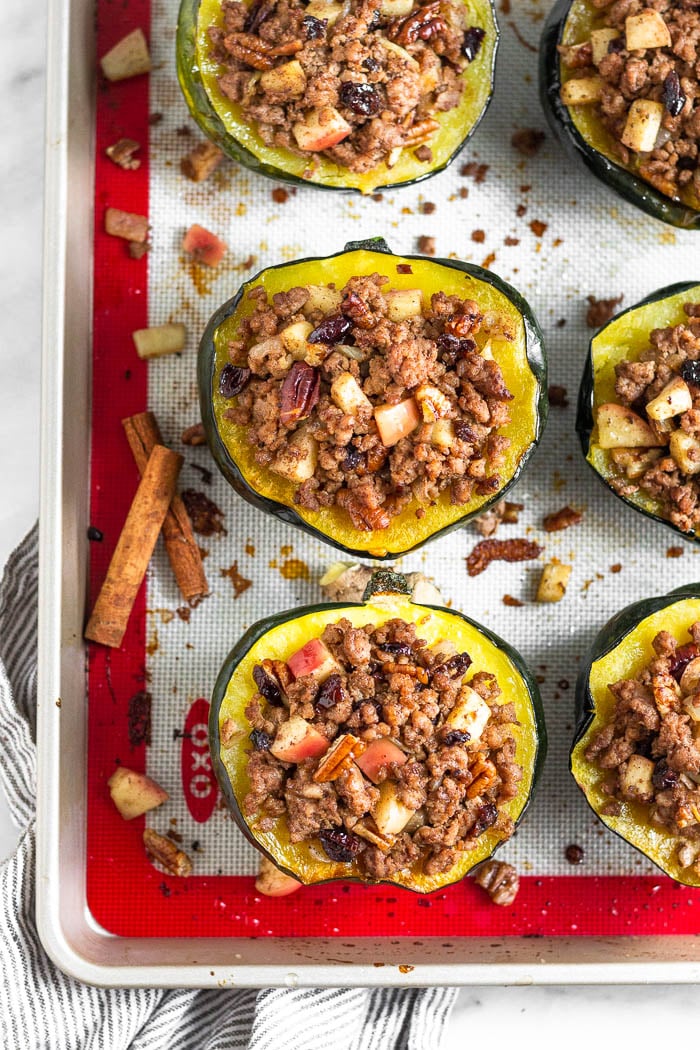 Sausage stuffed acorn squash on a baking sheet with a few pieces of the sausage and apple filling on the baking sheet as well. Two cinnamon sticks are also on the baking sheet.