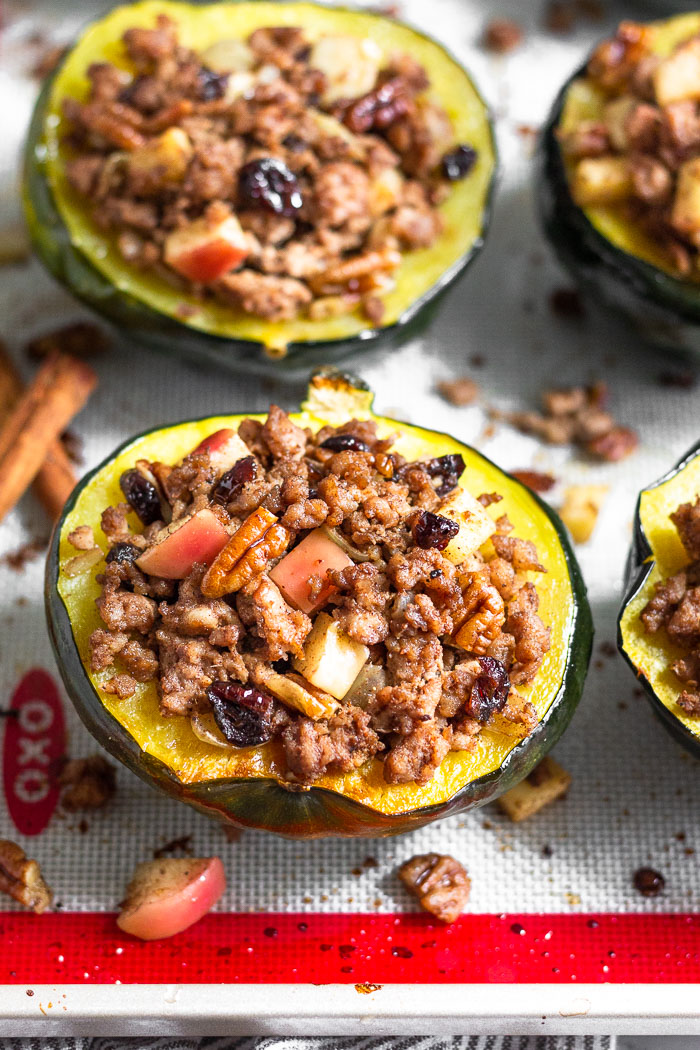 Baking sheet filled with stuffed acorn squash with the filling scattered around it as to make it look like they are overflowing. Two cinnamon sticks are on the baking sheet as well.