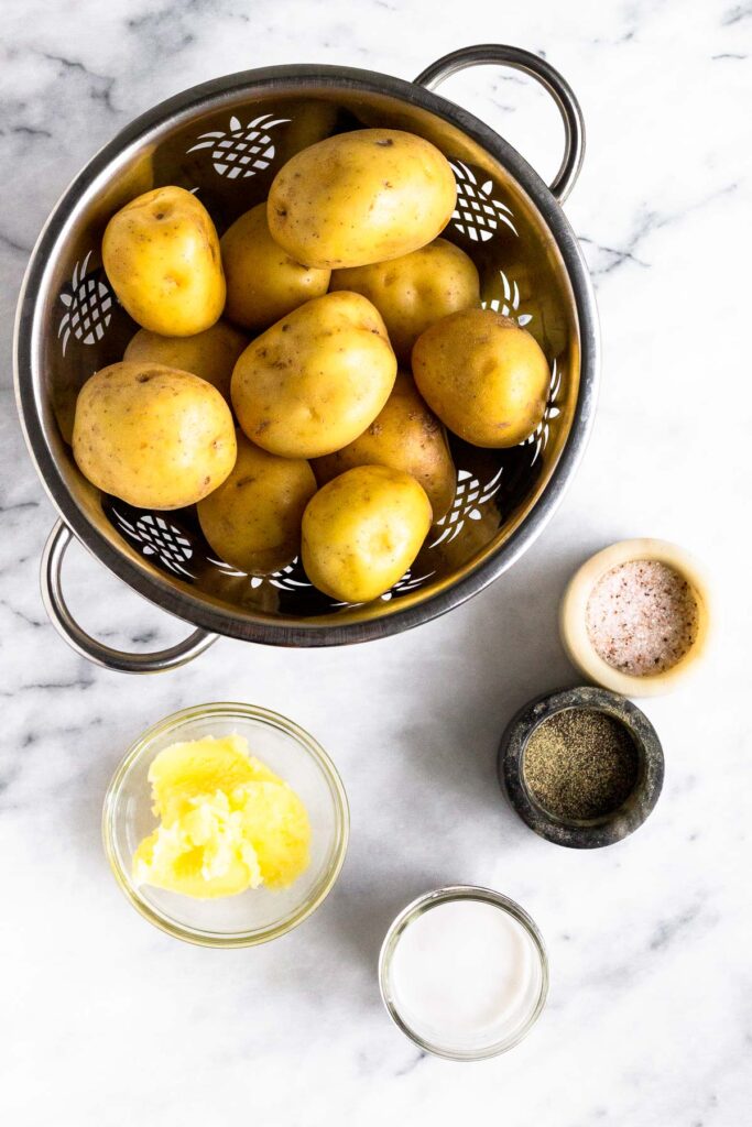 White counter with a colander of potatoes, containers of salt and pepper, a jar of coconut milk, and a bowl of ghee.
