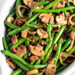 Green beans and mushrooms with bacon and fried onions in a white casserole dish. The dish is sitting on a striped towel with a lemon wedge next to it.