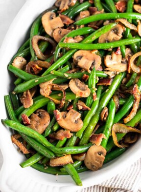 Green beans and mushrooms with bacon and fried onions in a white casserole dish. The dish is sitting on a striped towel with a lemon wedge next to it.